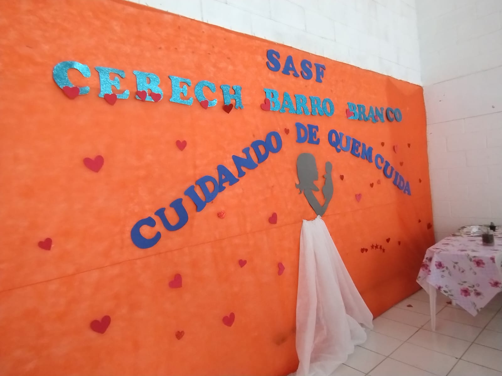 Na imagem, uma parede com um tecido laranja, escrito o nome da SASF e o nome da atividade, que é Cuidando de quem Cuida.